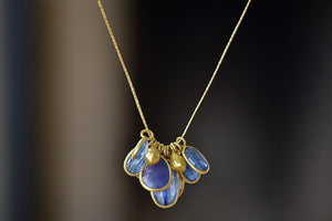 The Colette Cluster with Gold Drops, Kyanite and Tanzanite Necklace by Pippa Small is a large cluster of various sized bezel set and lightly faceted translucent stones in purple Tanzanite and blue Kyanite, accompanied by two gold beads, all in 18k yellow gold on a 20" golden waxed cotton cord form this necklace.