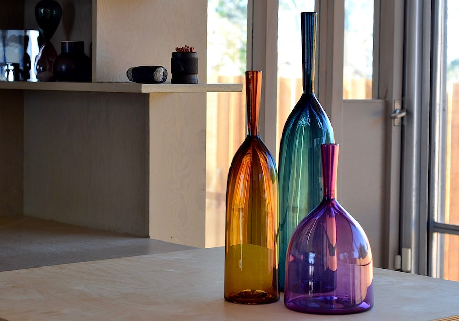 Angelic Bottles by Joe Cariati are mouth blown  Freeblown by glass master Joe Cariati in his Southern California studio, these bottle-form vases evoke the more mass produced mid-century ones that inspired them, large steel blue, small amber orange and wide ultra violet purple.