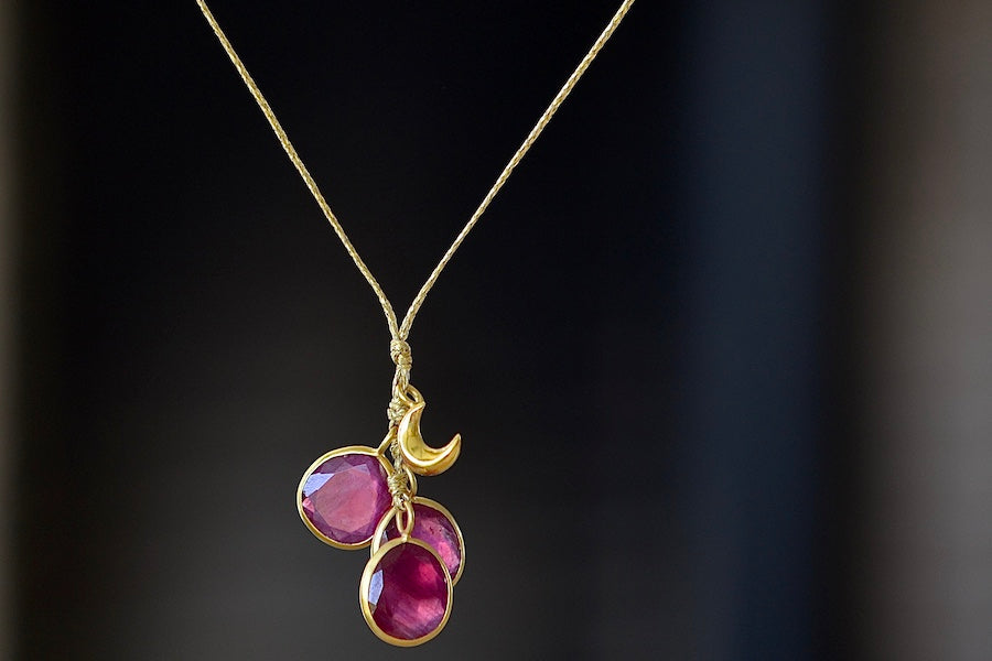 The Colette Set Cluster with Moon and Ruby Necklace is a  cluster of three bezel set and lightly faceted ruby stones, accompanied by a gold moon, all in 18k yellow gold on a 22" golden waxed cotton cord form this necklace. 