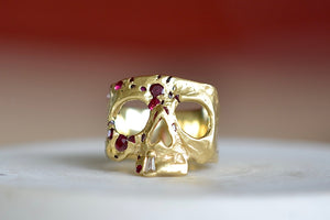 A Polly Wales Ruby Confetti Skull ring in 18k yellow gold is a square skull face ring with one diamond baguette snaggletooth and a scattering of red, pink and magenta rubies.