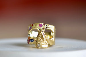 A Polly Wales Rainbow Confetti Skull ring in 18k yellow gold is a square skull face ring with one diamond baguette snaggletooth and a scattering of pink, magenta, red, orange, green, purple and blue sapphires.