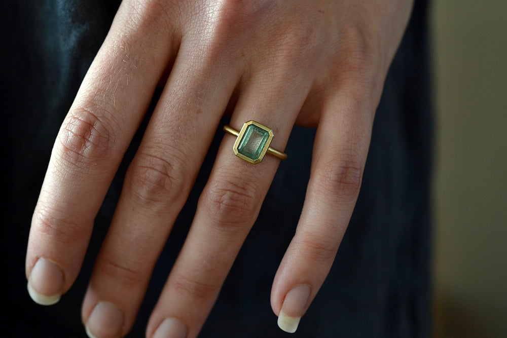 
            
                Load image into Gallery viewer, Wearing the The Elizabeth Street Jewelry Simple Emerald Band in size 6.5 is a bezel set (north/south) Colombian green emerald on a simple 14k satin yellow gold band. 
            
        