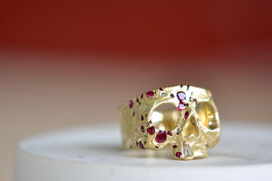 A Polly Wales Ruby Confetti Skull ring in 18k yellow gold is a square skull face ring with one diamond baguette snaggletooth and a scattering of red, pink and magenta rubies.