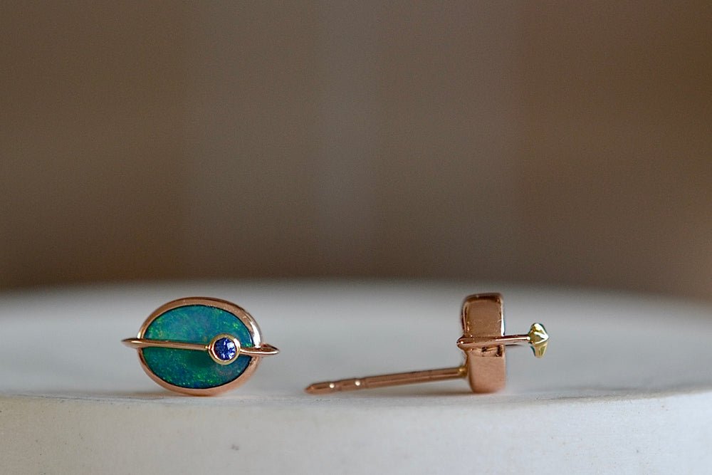 
            
                Load image into Gallery viewer, Side view of Small Planet Stud Earrings by Bibi Van Der Velden are comprised out of two bezel set opals with rings going through in rose gold of which one has a gold star and the other has a blue sapphire planet. 
            
        