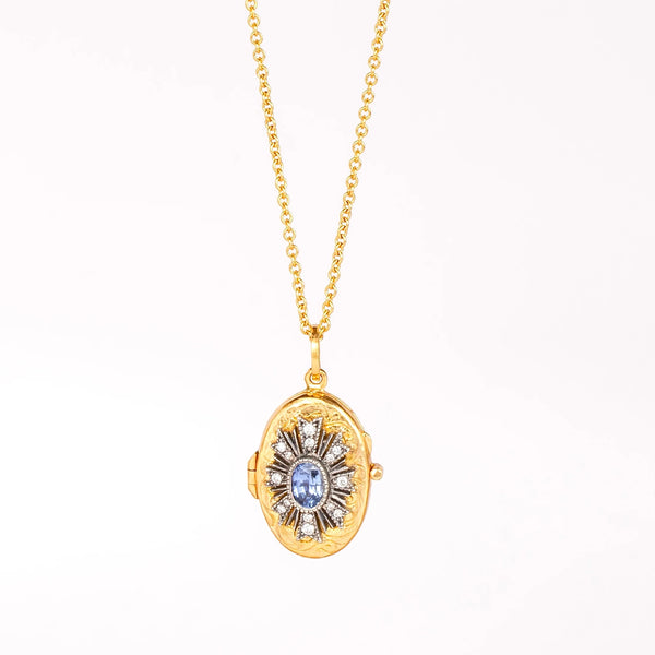 A Blue Sapphire Locket that opens by Arman Sarkisyan is an engraved oval disc in 22k with a blue center stone inside a flower motif made out of diamonds and hangs on a gold chain. Shown on white background.