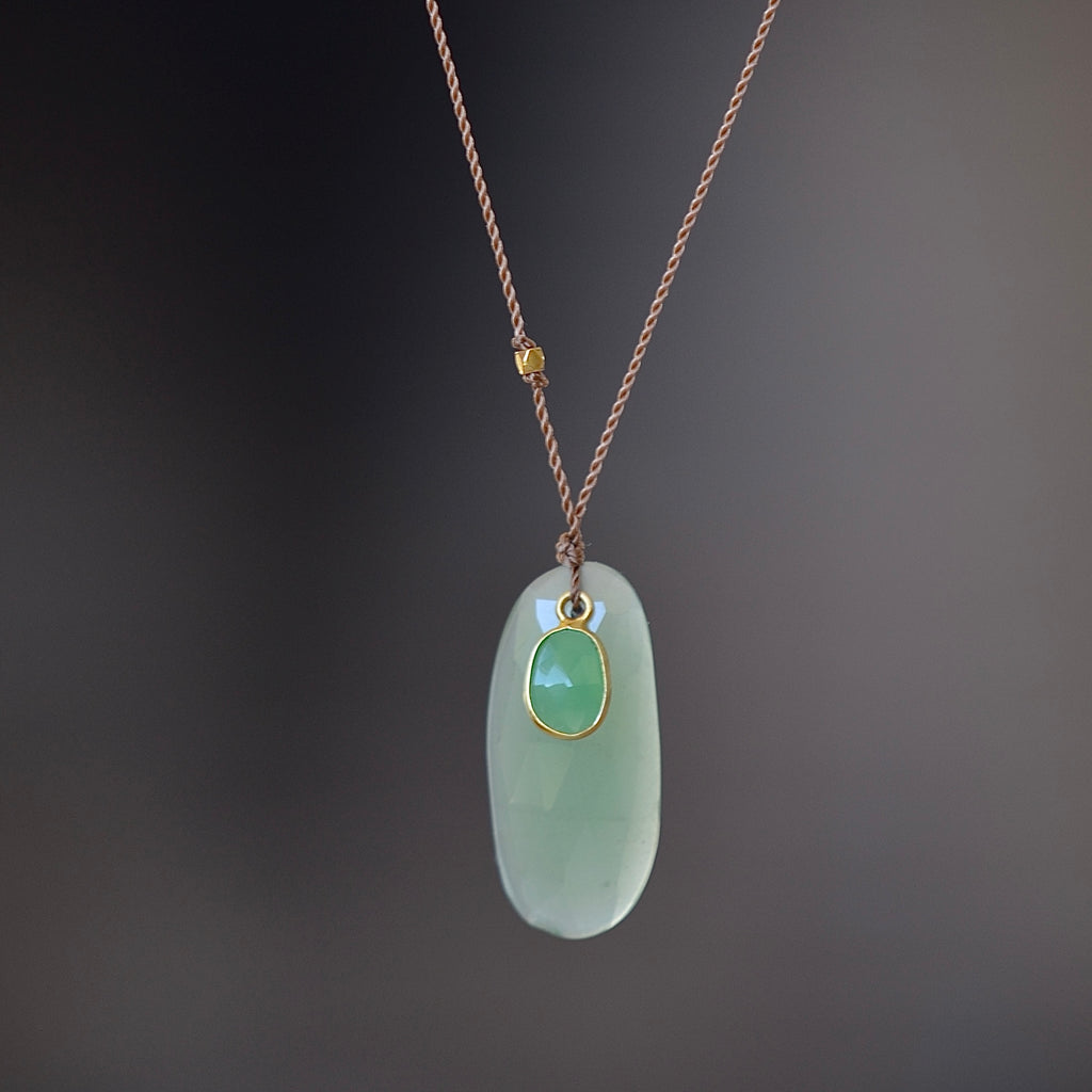 Chrysoprase and Sapphire Pendant Necklace by Margaret Solow is an organic necklace consisting of a smaller and bezel set green sapphire on top of a larger oval pale green raw chrysoprase that hangs on a cord with an 18k gold bead and has a clasp closure.