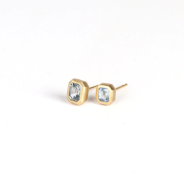 Bezel Set Asymmetrical Studs by Elizabeth Street Jewelry on white background. These are a pair of asymmetrical square/rectangle, soft purpley blue and bezel set sapphire stud earrings with post closure in 14k yellow gold. Handmade in Los Angeles.