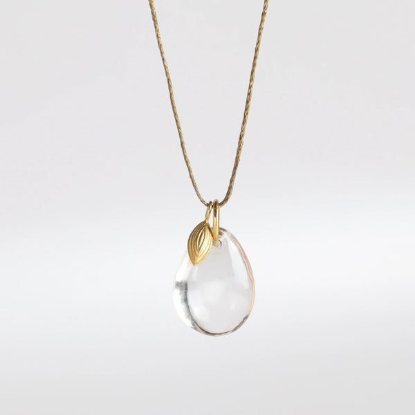 The Coconut Pendant with Seed Charm Necklace in Crystal  designed by Pippa Small is a One clear, transparent and smooth crystal stone with natural inclusions is drilled and hooked on to a gold loop, and accompanied by a bead in 18k yellow gold on a 24" golden waxed cotton cord form this necklace.