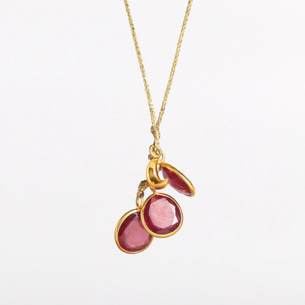 The Colette Set Cluster with Moon and Ruby Necklace is a  cluster of three bezel set and lightly faceted ruby stones, accompanied by a gold moon, all in 18k yellow gold on a 22" golden waxed cotton cord form this necklace. 