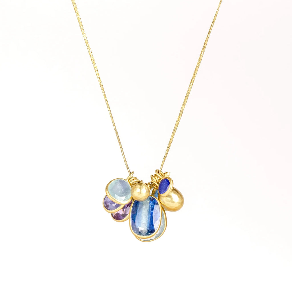 The Colette Mixed Blues Cluster Necklace by Pippa Small is A large cluster of nine various sized, bezel set, lightly faceted and translucent stones in the peacock color-way with aquamarine, amethyst, labradorite, kyanite, lapis and tanzanite, accompanied by a hammered gold seed and mini croissant beads in 18k gold and hanging on a 16" golden waxed cotton cord shown on white background.