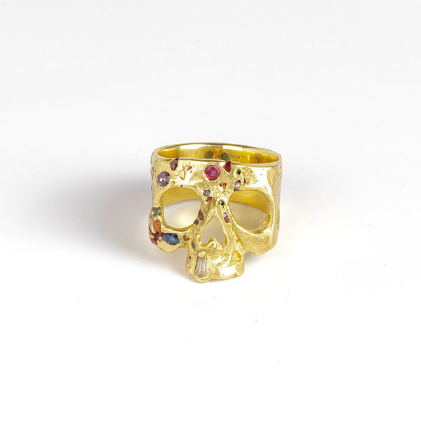 A Polly Wales Rainbow Confetti Skull ring in 18k yellow gold is a square skull face ring with one diamond baguette snaggletooth and a scattering of pink, magenta, red, orange, green, purple and blue sapphires on white background.