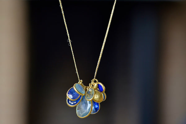 The Colette Mixed Blues Cluster Necklace by Pippa Small is A large cluster of nine various sized, bezel set, lightly faceted and translucent stones in the peacock color-way with aquamarine, amethyst, labradorite, kyanite, lapis and tanzanite, accompanied by a hammered gold seed and mini croissant beads in 18k gold and hanging on a 16" golden waxed cotton cord.