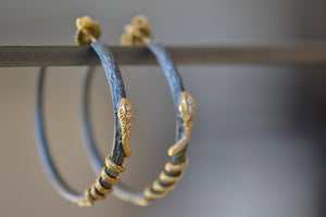 The Snake Hoop Earrings by Arman Sarkyssian are two 22k gold snakes with diamond pavé details that are entwined on an oxidized silver hoop textured like a tree branch in oxidized sterling silver with a post backing.