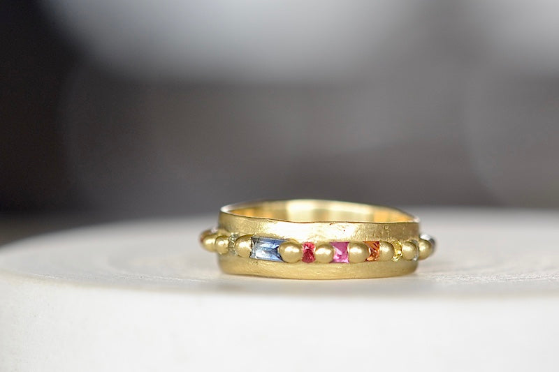 
            
                Load image into Gallery viewer, The Wide Harlequin Nina Ring is a wide and organically shaped pinch band in 18k recycled yellow gold is encrusted with mixed cut Rainbow Sapphires in pink, blue, yellow, orange and green sapphires around the circumference. Recycled gold. Cast in Place. Cast not set. Handmade in Los Angeles.
            
        