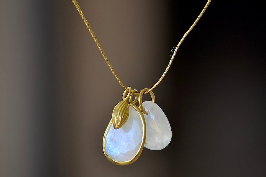 The Colette Drill Pendant with Seed Charm in Moonstone is a cluster of two milky white rainbow moonstones of which one is bezel set and lightly faceted and the other is smooth and round. These are accompanied by a sun bead, all in 18k yellow gold on a 20" golden waxed cotton cord form this necklace. Medium shot.