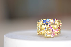 Encrusted River Skull Ring is a Polly Wales' classic squared 18k yellow gold small face skull ring, encrusted with gradated rainbow sapphires in her pebbles on a  river style and with one white diamond baguette snaggletooth. Recycled gold. Cast in Place. Cast not set size 7.5. Handmade in Los Angeles.