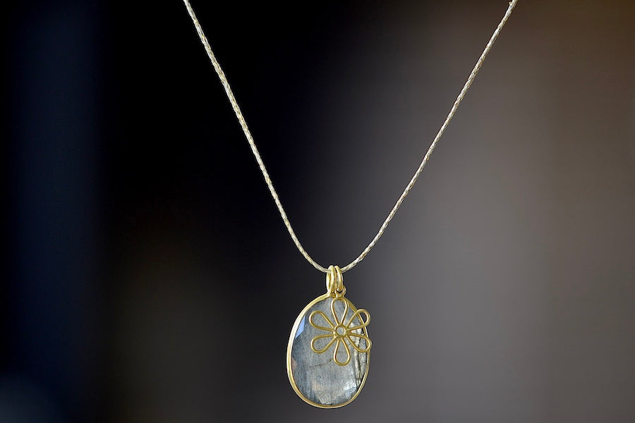 The Colette Pendant in Labradorite by Pippa Small is made out of one light grey, translucent, iridescent, lightly and faceted labradorite stone which is bezel set and accompanied by a flower charm, all in 18k yellow gold on a 26" golden waxed cotton cord form this necklace.