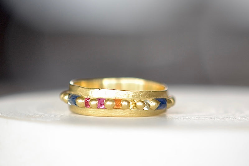 The Wide Harlequin Nina Ring is a wide and organically shaped pinch band in 18k recycled yellow gold is encrusted with mixed shape Rainbow Sapphires in pink, blue, yellow, orange and green sapphires around the circumference. Recycled gold. Cast in Place. Cast not set. Handmade in Los Angeles.
