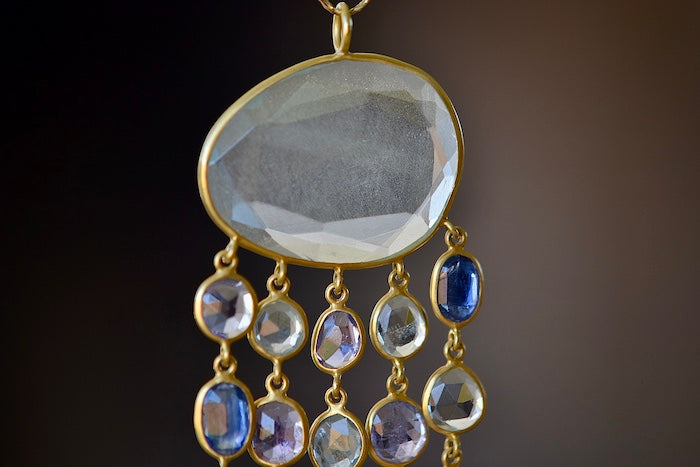 Raincloud Pendant Necklace by Pippa Small is a Large, faceted and transparent aquamarine with five rows of five stones each in blue to light purple hued tanzanite, aquamarine and kyanite bezel set stones in 18k yellow gold w extra long golden waxed cotton cord and accent bead for draping over head and no clasp.
