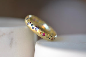 
            
                Load image into Gallery viewer, Tilted Polly Wales Classic Rainbow Confetti Band Ring in 18k recycled yellow gold with green, yellow, orange, blue, purple and pink sapphires and cast not set. Size 7. Available at OK.
            
        