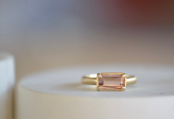 Elizabeth Street Jewelry Eagle Claw Bar ring in light pink peach tourmaline , clear and translucent, tourmaline is a radiant cut pink tourmaline in a four prong eagle claw east/west bezel setting on a 14k satin 3mm yellow gold band size 7..