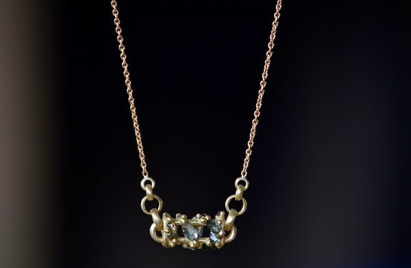 Close up of Horizontal Fontaine Bar Necklace in Green by Polly Wales is An oval bar in 18K yellow gold has a vine of encrusted inverted sapphires in green, gray and black that have been cast and are accompanied by matte gold dots. The bar hangs on a beautiful gold chain. Recycled Gold. Handmade in Los Angeles. Cast not set.