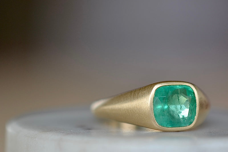 A signet ring in green emerald by Elizabeth street jewelry is a vivid cushion cut and lightly faceted Colombian emerald on a tapered band in 18k satin finish yellow gold. Handmade in Los Angeles.