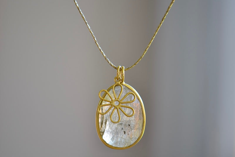 The Colette Pendant in Labradorite by Pippa Small is made out of one light grey, translucent, iridescent, lightly and faceted labradorite stone which is bezel set and accompanied by a flower charm, all in 18k yellow gold on a 26" golden waxed cotton cord form this necklace.