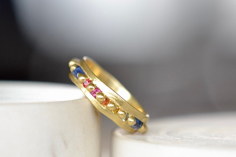The Wide Harlequin Nina Ring is a wide and organically shaped pinch band in 18k recycled yellow gold is encrusted with mixed shape Rainbow Sapphires in pink, blue, yellow, orange and green sapphires around the circumference. Recycled gold. Cast in Place. Cast not set. Handmade in Los Angeles.