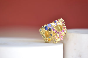 
            
                Load image into Gallery viewer, Encrusted River Skull Ring is a Polly Wales&amp;#39; classic squared 18k yellow gold small face skull ring, encrusted with gradated rainbow sapphires in blue, pink green, yellow, orange and lilac in her pebbles on a  river style and with one white diamond baguette snaggletooth. Recycled gold. Cast in Place. Cast not set size 7.5. Handmade in Los Angeles.
            
        