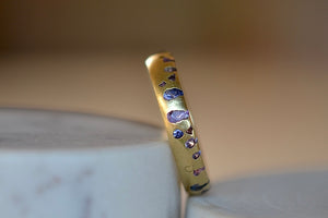 Tilted view of the Purple and Blue Sapphire Band Ring by Polly Wales is a narrow 18k yellow gold band with speckled purple, pink, blue to navy and black sapphires around the circumference for a beautiful confetti-like appearance. Recycled gold. Cast not set.