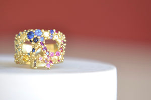 
            
                Load image into Gallery viewer, Encrusted River Skull Ring is a Polly Wales&amp;#39; classic squared 18k yellow gold small face skull ring, encrusted with gradated rainbow sapphires in blue, pink green, yellow, orange and lilac inher pebbles on a river style and with one white diamond baguette snaggletooth. Recycled gold. Cast in Place. Cast not set size 7.5. Handmade in Los Angeles.
            
        