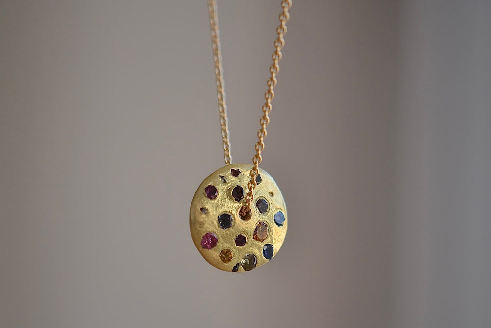 Polly Wales Spinning Disc Pendant Necklace in 18K yellow gold with scattered rainbow sapphires in orange, yellow, pink, blue, purple and lilac hangs on a beautiful chain 22" cast in place and cast not set in recycled gold.