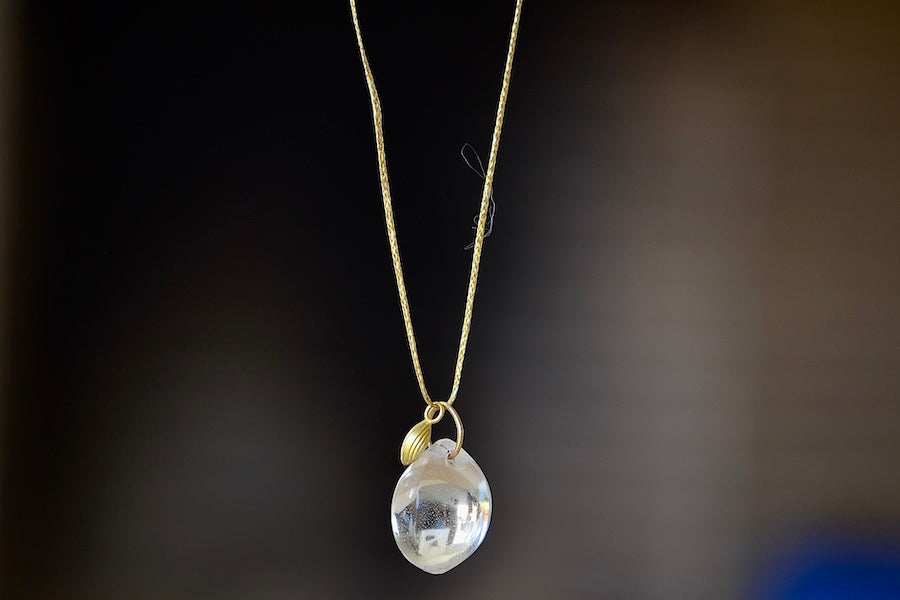 The Coconut Pendant with Seed Charm Necklace in Crystal  designed by Pippa Small is a One clear, transparent and smooth crystal stone with natural inclusions is drilled and hooked on to a gold loop, and accompanied by a bead in 18k yellow gold on a 24" golden waxed cotton cord form this necklace.