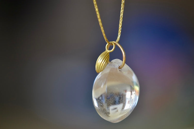 The Coconut Pendant with Seed Charm Necklace in Crystal  designed by Pippa Small is a One clear, transparent and smooth crystal stone with natural inclusions is drilled and hooked on to a gold loop, and accompanied by a bead in 18k yellow gold on a 24" golden waxed cotton cord form this necklace.
