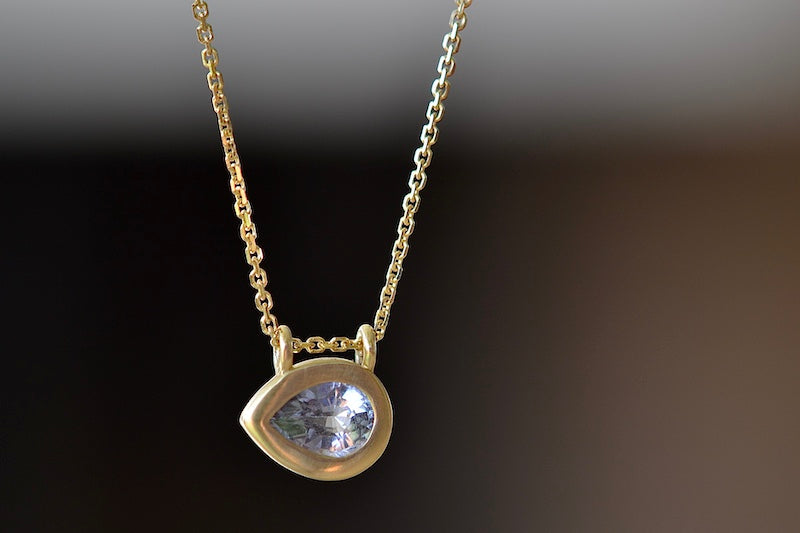 Duo Bale Sapphire Teardrop Pendant Necklace by Elizabeth Street is a bezel set teardrop shaped purple to lilac or violet sapphire set on its side (east/west) in a double bale and on a 14k yellow gold chain. Perfect for layering or as a solo piece. Handmade in Los Angeles.